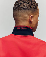 A person with short curly hair is shown from behind wearing the Psycho Bunny MENS JUDE PIQUE POLO - MT0100012, featuring a black collar, red "PSYCHO BUNNY" embroidery, the signature Bunny detail, and soft Pima cotton fabric.