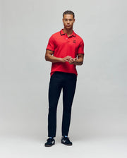 A man stands against a plain background wearing the Psycho Bunny MENS JUDE PIQUE POLO - MT0100012 in red. He faces forward with his hands together and looks aside, showing off the signature embroidered Bunny logo.