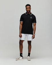 A man stands facing forward in a Psycho Bunny MENS JUDE PIQUE POLO - MT0100012, paired with white shorts, black socks, and white sneakers, against a plain light gray background.