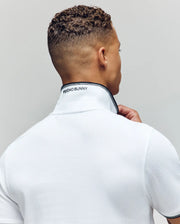 A person with short curly hair, seen from behind, wears the Psycho Bunny MENS JUDE PIQUE POLO (MT0100012) in white Pima cotton, displaying "PSYCHO BUNNY" text and an embroidered Bunny on the collar as they turn and hold it up.