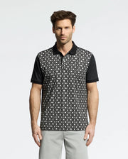A man models the Psycho Bunny MENS BECKETT PIQUE POLO - MT0100022, featuring a black and white geometric front with solid black sleeves and collar. Against a white backdrop, he pairs it with light gray pants, exuding casual elegance.