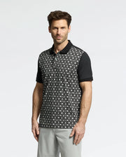 A man is wearing the Psycho Bunny MENS BECKETT PIQUE POLO - MT0100022, featuring a black and white golf-ball pattern with short sleeves, paired with gray pants, against a plain white background.