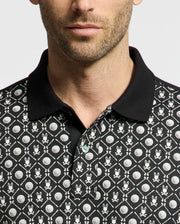 A man wears the Psycho Bunny MENS BECKETT PIQUE POLO, MT0100022, featuring a black Pima cotton base with playful white geometric patterns like golf-ball-style circles and abstract bunny motifs. It includes a black collar contrasting against its detailed design on a plain white background.