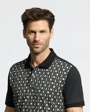 A man with short brown hair and a trimmed beard stands against a white background, wearing the Psycho Bunny MENS BECKETT PIQUE POLO - MT0100022. The Pima cotton polo with geometric patterns and floral designs, along with its four-way stretch, enhances his slight smile perfectly.