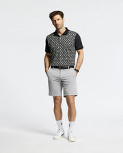 A man stands with hands in his pockets, wearing a Psycho Bunny MENS BECKETT PIQUE POLO (MT0100022), light gray shorts, white socks, and white sneakers against a plain light background.