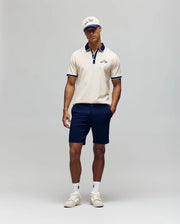 A man stands against a plain background wearing the Psycho Bunny MENS MATTEO PIQUE POLO - MT0100024, paired with navy shorts, white sneakers, a white cap, and sunglasses. His hands are in his pockets and he looks relaxed.