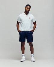 A man stands against a plain background wearing a white Psycho Bunny Mens Mischief Bunny Pique Polo (MT0100025) with a small red emblem, navy shorts, white socks with blue stripes, and white high-top sneakers. He looks relaxed with hands in pockets.