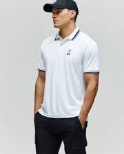 A man wearing the Psycho Bunny MENS CHRIS PERFORMANCE PIQUE POLO - MT0100038, featuring a white fabric with blue-striped collar and cuffs, pairs it with black cargo pants and a black baseball cap, standing with hands in pockets and looking to the side.