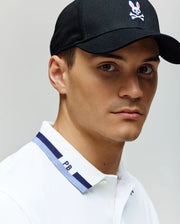 A young man wearing a Psycho Bunny black cap and the MENS CHRIS PERFORMANCE PIQUE POLO - MT0100038, a white slim fit polo with blue trim and "PB" on the collar, looks directly at the camera with a neutral expression.