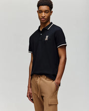 A young man stands against a plain background wearing the Psycho Bunny MENS WADE PIQUE POLO - MT0100044 in black with white trim and a chest logo, paired with tan cargo pants.