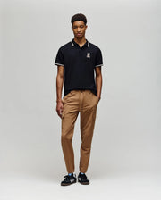 A person stands against a plain background wearing the Psycho Bunny MENS WADE PIQUE POLO - MT0100044 in classic black with a white logo, paired with tan drawstring pants and black sneakers featuring white stripes.