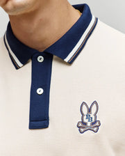 A close-up shows a person wearing the Psycho Bunny MENS WADE PIQUE POLO (MT0100044), a colorblock style with a navy collar, buttons, and an embroidered bunny logo above crossed bones plus "P B" on the left chest.