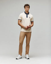 A man with curly hair and a neutral expression stands against a plain background wearing the Psycho Bunny MENS WADE PIQUE POLO - MT0100044, tan pants, and white sneakers. His hands are loosely clasped.