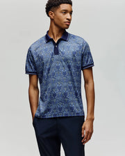 A young man wears the Psycho Bunny MENS REEVES ALL OVER BUNNY SPORT POLO (MT0100053), a blue patterned, moisture-wicking polo with a dark collar, paired with dark pants, standing against a plain light gray background.