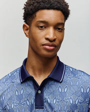 A young man with short curly hair models the Psycho Bunny MENS REEVES ALL OVER BUNNY SPORT POLO (MT0100053), featuring a breathable fabric, subtle blue pattern, and dark blue collar against a plain light background.