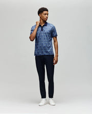 A man stands against a plain light gray background, wearing the Psycho Bunny MENS REEVES ALL OVER BUNNY SPORT POLO (MT0100053), dark navy pants, and white sneakers. He is turned slightly and holds his shirt collar with one hand.
