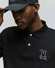 A man in a Psycho Bunny MENS CYRUS PIQUE POLO - MT0100056, featuring soft black Pima cotton and a white bunny logo, adjusts his matching black cap while looking down.