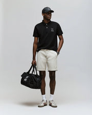 A man wearing a Psycho Bunny MENS CYRUS PIQUE POLO - MT0100056 in black, beige shorts, white socks, sneakers, a black cap, and holding a black duffel bag stands against a plain light background.