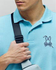 A person wears the Psycho Bunny MENS CYRUS PIQUE POLO - MT0100056 in light blue with the signature embroidered bunny logo, holding a black strap. Only their lower face, chest, and hand are visible in the cropped image.