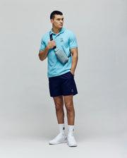 A man stands against a plain background wearing Psycho Bunny's MENS CYRUS PIQUE POLO - MT0100056 in light blue, navy shorts, white sneakers, and a light gray crossbody bag. His right hand is in his shorts pocket; left holds the bag’s strap.