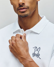 A man wearing the Psycho Bunny MENS CYRUS PIQUE POLO (MT0100056) with white Pima cotton, large bunny embroidery and crossed bones on the chest, holding the collar. Only his lower face and upper torso are visible.