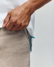 A close-up shows a hand holding the hem of the Psycho Bunny MENS CYRUS PIQUE POLO - MT0100056, white Pima cotton with a small blue tag. The person wears light beige pants against a plain, neutral background.