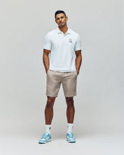 A man stands against a plain background wearing the Psycho Bunny MENS CYRUS PIQUE POLO - MT0100056 in white, beige shorts, white socks, and light blue sneakers. His hands are in his pockets as he looks confidently at the camera.