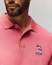 A man wears the Psycho Bunny MENS SONNY PIQUE POLO—crafted from soft Pima cotton—featuring unique chest embroidery: a skull and crossbones under palm trees with a small green ball.