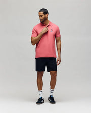 A man with short hair and a beard stands against a plain background, touching his collar as he looks down. He wears Psycho Bunny's MENS SONNY PIQUE POLO - MT0100094 in pink, black shorts, striped white socks, and black sneakers.