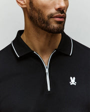 A man with a short beard wears the Psycho Bunny MENS DANE ZIP PIQUE POLO (MT0100103), a black collared zip-up shirt with white trim, sleek zip placket, and an embroidered Bunny logo on the chest. Image is cropped at nose and shoulders.
