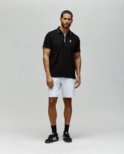 A man stands against a plain background wearing the Psycho Bunny MENS DANE ZIP PIQUE POLO - MT0100103 in black with white trim, paired with white knee-length shorts, black crew socks, and black sneakers.