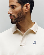 A man with short dark hair and a trimmed beard wears the Psycho Bunny Mens Henry Oversized Fit Polo (MT0100104) in cream, featuring a small blue bunny and crossbones logo, looking to his left against a neutral background.