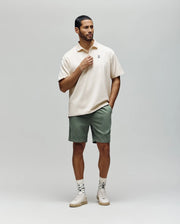 A man stands against a plain background wearing the Psycho Bunny MENS HENRY OVERSIZED FIT POLO - MT0100104, sage green shorts, patterned white socks, and white sneakers. One hand touches his collar while the other rests in his pocket.