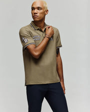 A man with short, bleached blonde hair wears the Psycho Bunny MENS WADE PATCH PIQUE POLO SHIRT (MT0100109) in khaki, featuring an embroidered Bunny logo and "Psycho Bunny" text, paired with navy pants against a plain light background.