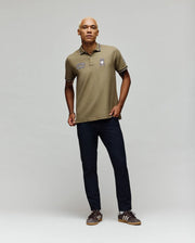 A person with short blond hair stands in a studio wearing the Psycho Bunny MENS WADE PATCH PIQUE POLO SHIRT (MT0100109), dark pants, and brown sneakers against a plain light gray background.