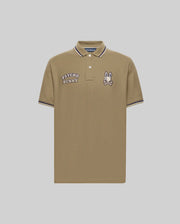 The Psycho Bunny Men's Wade Patch Pique Polo Shirt (MT0100109) is made from khaki Pima cotton with white buttons, "PSYCHO BUNNY" text, an embroidered Bunny skull logo patch on the chest, and white/navy striped collar and sleeve trim.