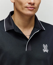 A person wears the Psycho Bunny MENS JAYDEN SPORT POLO - MT0100111 in black with white trim and an embroidered bunny logo on the left chest; moisture-wicking fabric ensures comfort, showing only the lower face and upper torso.