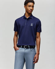 A man wears the Psycho Bunny MENS JAYDEN SPORT POLO - MT0100111 in dark navy with a white logo and athletic fit, paired with light gray trousers, standing against a plain light background with one hand in his pocket.
