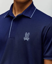 A man wears the Psycho Bunny MENS JAYDEN SPORT POLO - MT0100111 in navy with light blue collar piping and a white feather and bones logo, showcasing its athletic fit on his upper torso and part of his face.