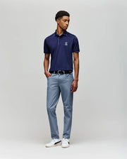 A man stands against a plain light background, wearing a Psycho Bunny MENS JAYDEN SPORT POLO - MT0100111 with moisture-wicking fabric, paired with light blue trousers, a black belt, and white sneakers. He looks slightly left with his left hand in his pocket.