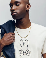 A man in the Psycho Bunny MENS SETH GRAPHIC TEE (MT0200005), featuring a bold skull and crossbones, holds a dark jacket over his shoulder and gazes to the side against a plain background.