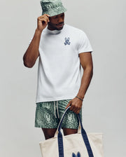 A man wears the Psycho Bunny MENS DEREK CHEST GRAPHIC TEE (MT0200009), green patterned shorts, a matching bucket hat, and carries a beige tote bag with navy handles and Bunny logo, standing against a plain light background.