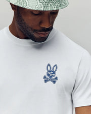 A man wearing a light patterned bucket hat and the Psycho Bunny MENS DEREK CHEST GRAPHIC TEE (MT0200009), a white regular fit t-shirt with a blue bunny ears and crossed bones logo, looking downwards.