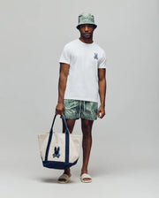 A man stands against a plain background wearing a light green bucket hat, green patterned shorts, beige sandals, and holding a large beige and navy tote bag with a bunny print. He sports Psycho Bunny’s MENS DEREK CHEST GRAPHIC TEE - MT0200009.
