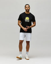 A man stands against a plain background wearing Psycho Bunny's MENS PETE GRAPHIC TEE (MT0200012) in black Pima cotton, paired with white shorts, white tennis socks featuring black stripes, and white sneakers with green accents.