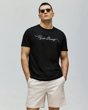 A man wearing sunglasses, the Psycho Bunny MENS CYRUS WORDMARK GRAPHIC TEE - MT0200018 in black pima cotton, and light beige shorts stands with his hands in his pockets against a plain, light background.