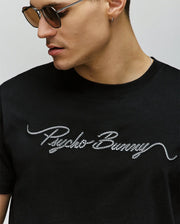 A man wearing sunglasses and a black Psycho Bunny MENS CYRUS WORDMARK GRAPHIC TEE (MT0200018) with white script "Psycho Bunny" embroidered across the chest.