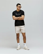 A man in sunglasses, beige shorts, white socks and sneakers wears the Psycho Bunny MENS CYRUS WORDMARK GRAPHIC TEE - MT0200018 (black with white script), standing with arms crossed against a light background.