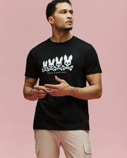 A man models the Psycho Bunny MENS ARCHIVE GRADIANT BUNNY GRAPHIC TEE (MT0200029), a bold black shirt with four Bunny skull-and-crossbones designs and "GOLD SINCE 2005" text, standing against a pink background, gesturing as he looks aside.