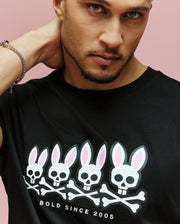 A man poses against a pink background wearing the Psycho Bunny MENS ARCHIVE GRADIANT BUNNY GRAPHIC TEE (MT0200029), a black shirt with four cartoon skulls, pink bunny ears and crossbones, and the "BOLD SINCE 2005" text.
